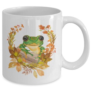 May include: White ceramic coffee mug featuring a green frog perched on a log, surrounded by autumn leaves and flowers. The frog has large, black eyes and a yellow belly.