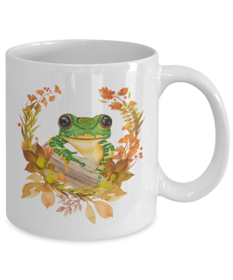 May include: White ceramic coffee mug featuring a green frog perched on a log, surrounded by autumn leaves and flowers. The frog has large, black eyes and a yellow belly.