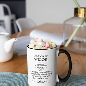 May include: White ceramic mug with a black handle and rim, filled with colorful marshmallows. The mug features the text "Potion of Vigor" with additional text below. The mug is on a wooden table.