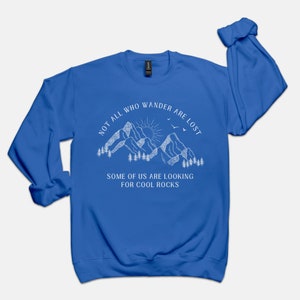 May include: Royal blue long-sleeve sweatshirt with a white graphic of mountains, a sun, and birds. The text reads "Not all who wander are lost. Some of us are looking for cool rocks."