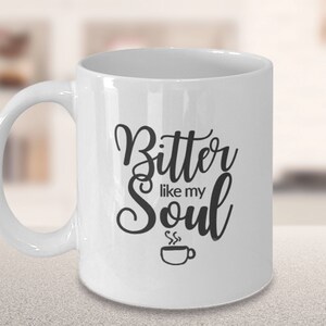 Op de afbeelding: Witte keramische koffiemok met zwarte tekst die "Bitter like my Soul" luidt en een klein zwart koffiekopje-icoon met stoom die eruit komt.