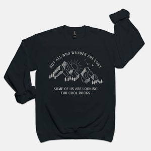 May include: Black sweatshirt with a white graphic of mountains, a sun, and birds. The text reads "Not all who wander are lost. Some of us are looking for cool rocks."