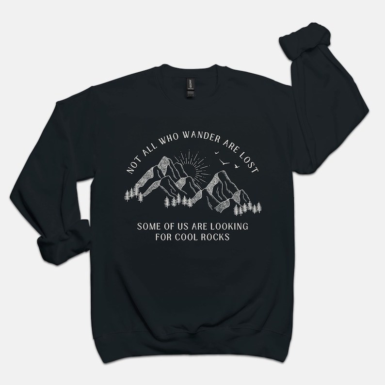 May include: Black sweatshirt with a white graphic of mountains, a sun, and birds. The text reads "Not all who wander are lost. Some of us are looking for cool rocks."