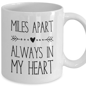 Pu&ograve; includere: Tazza da caff&egrave; in ceramica bianca con testo nero che recita "Miles Apart Always In My Heart" con un piccolo cuore e frecce su entrambi i lati del cuore.