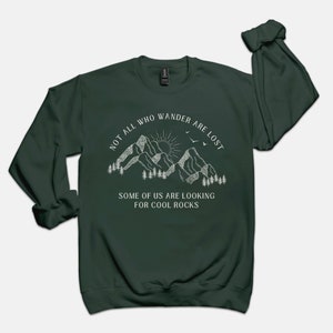 May include: A dark green sweatshirt with a white graphic of mountains, a sun, and birds. The text reads "Not all who wander are lost. Some of us are looking for cool rocks."