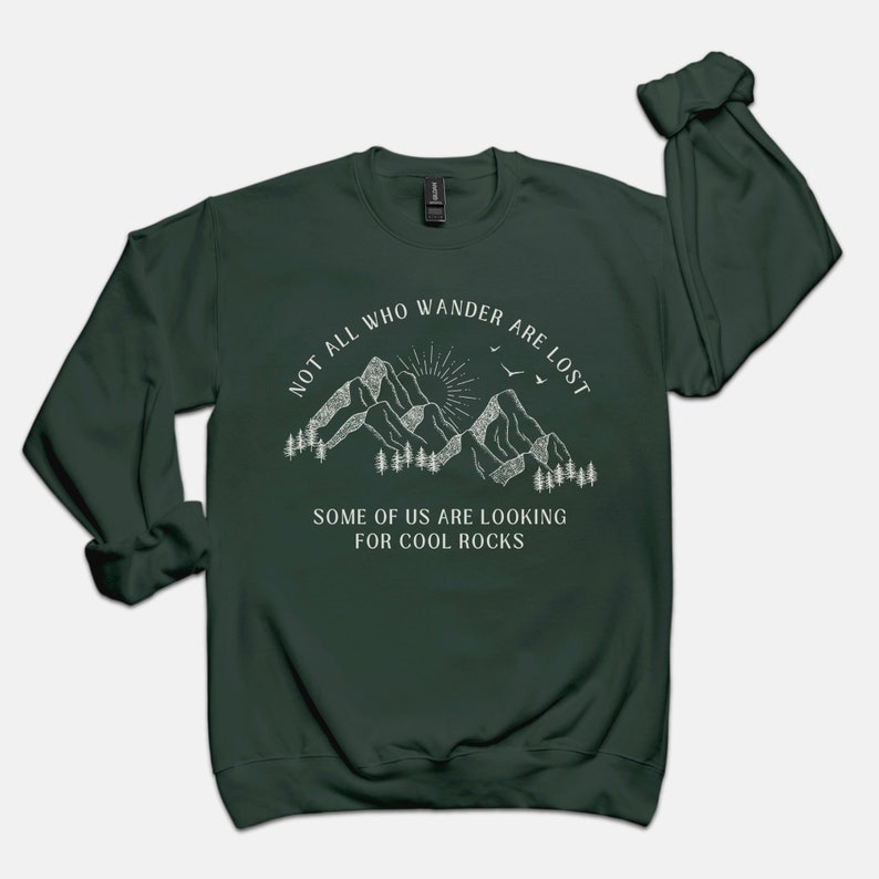 May include: A dark green sweatshirt with a white graphic of mountains, a sun, and birds. The text reads "Not all who wander are lost. Some of us are looking for cool rocks."