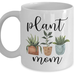 以下が含まれることがあります： 白いセラミック製のマグカップに、黒い文字で「plant mom」と書いてあります。テキストの上に、鉢植えの植物のウォーターカラーイラストが3つあります。