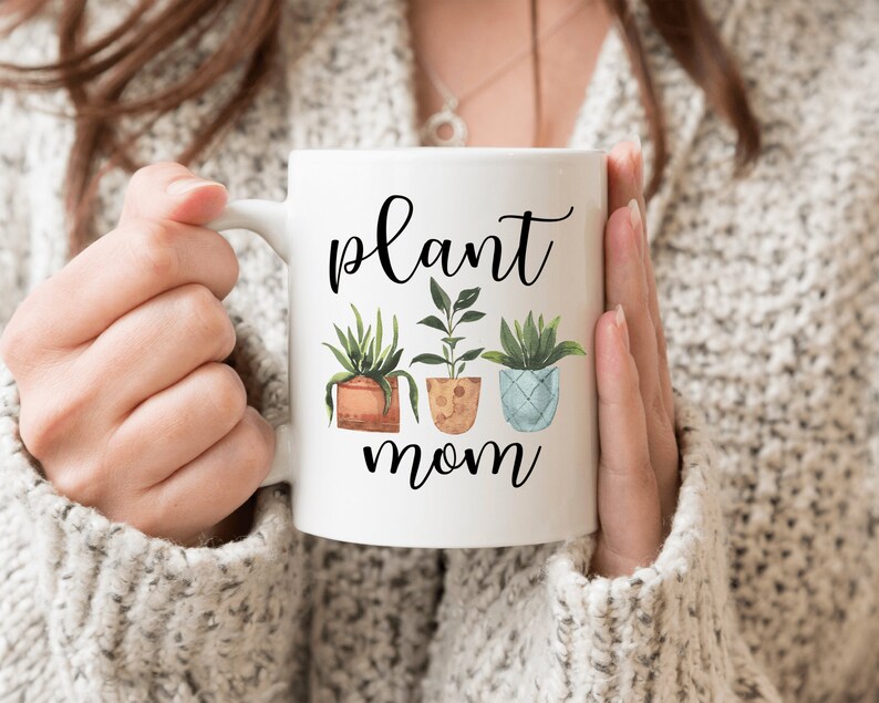 以下が含まれることがあります： 白いセラミック製のマグカップに、黒い筆記体で「plant mom」と書いてあり、3つの鉢植えの植物のグラフィックが描かれています。植物は緑、茶色、青です。