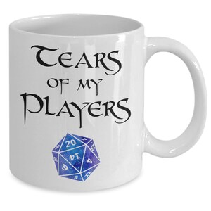 Op de afbeelding: Witte keramische koffiemok met de tekst "Tears of my Players" en een blauwe en paarse 20-zijdige dobbelsteen.