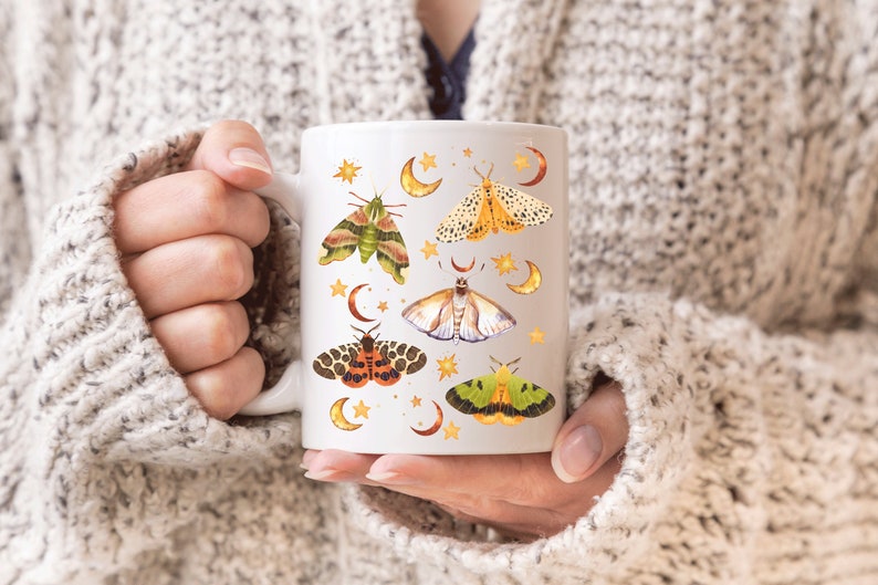 May include: White ceramic mug with a design of colorful moths and crescent moons. The moths are illustrated in a vintage style with intricate details.
