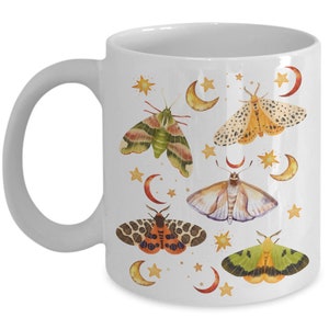 May include: White ceramic mug with a colorful moth and crescent moon design. The moths are illustrated in shades of green, orange, yellow, and brown. The crescent moons are yellow and the stars are gold.