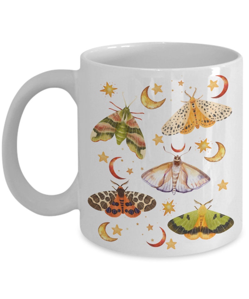 May include: White ceramic mug with a colorful moth and crescent moon design. The moths are illustrated in shades of green, orange, yellow, and brown. The crescent moons are yellow and the stars are gold.