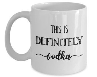 May include: White ceramic coffee mug with black text that reads "This is definitely vodka".
