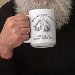 K&ouml;nnte beinhalten: Eine wei&szlig;e Keramiktasse mit einer Schwarzwei&szlig;illustration von Bergen und B&auml;umen. Der Text auf der Tasse lautet "Not all who wander are lost. Some of us are looking for cool rocks."