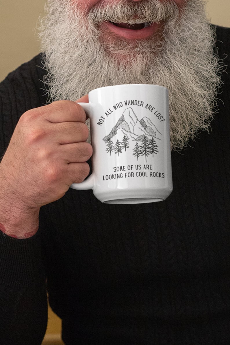 K&ouml;nnte beinhalten: Eine wei&szlig;e Keramiktasse mit einer Schwarzwei&szlig;illustration von Bergen und B&auml;umen. Der Text auf der Tasse lautet "Not all who wander are lost. Some of us are looking for cool rocks."