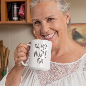 May include: A white ceramic mug with a black inscription that reads "Badass Nurse" and a small floral design. The mug is being held by a person in a white shirt with lace detailing.