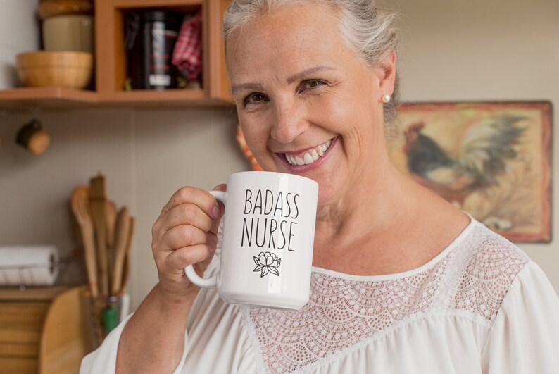 May include: A white ceramic mug with a black inscription that reads "Badass Nurse" and a small floral design. The mug is being held by a person in a white shirt with lace detailing.