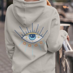May include: A light gray hooded sweatshirt with a blue eye design on the back. The eye has a white pupil, a blue iris, and a yellow crescent moon above it. The eye is surrounded by a yellow and orange sunburst.