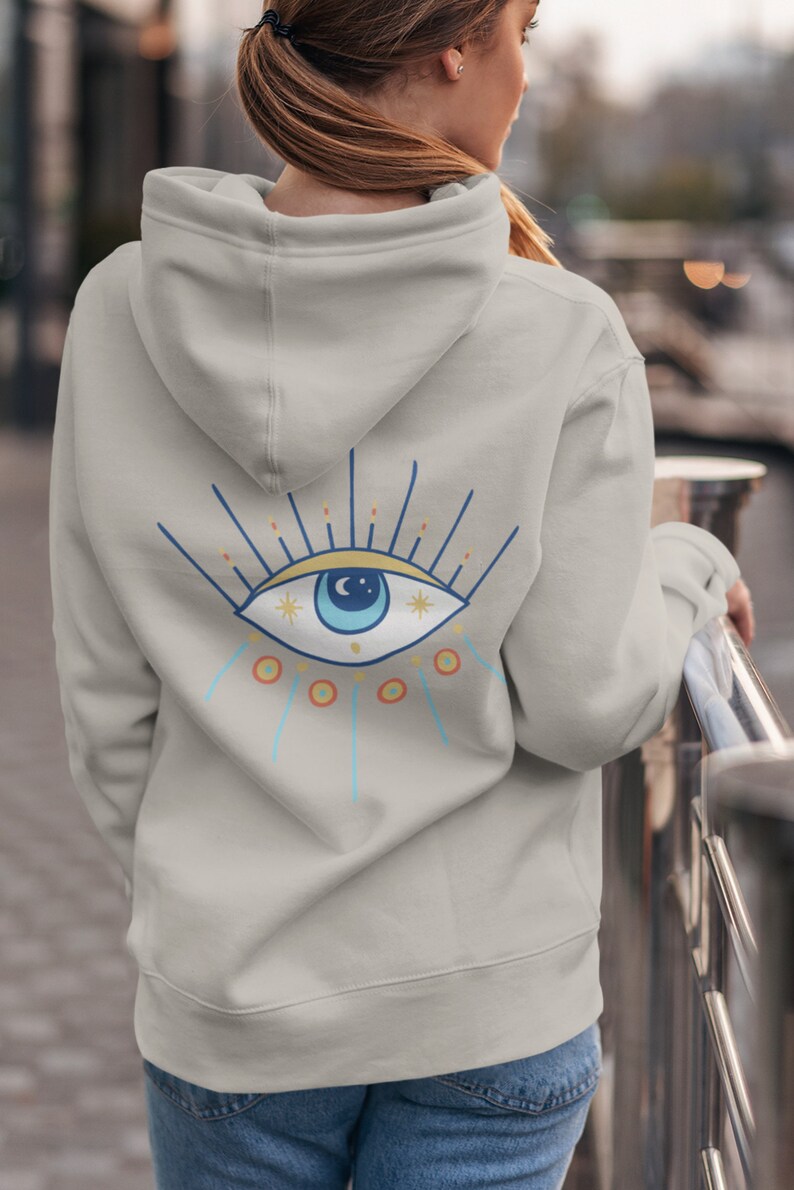 May include: A light gray hooded sweatshirt with a blue eye design on the back. The eye has a white pupil, a blue iris, and a yellow crescent moon above it. The eye is surrounded by a yellow and orange sunburst.