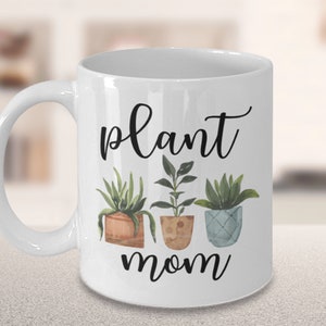 以下が含まれることがあります： 白いセラミック製のマグカップに、黒い筆記体で「plant mom」と書いてあり、緑の葉のついた鉢植えの植物が3つ描かれています。