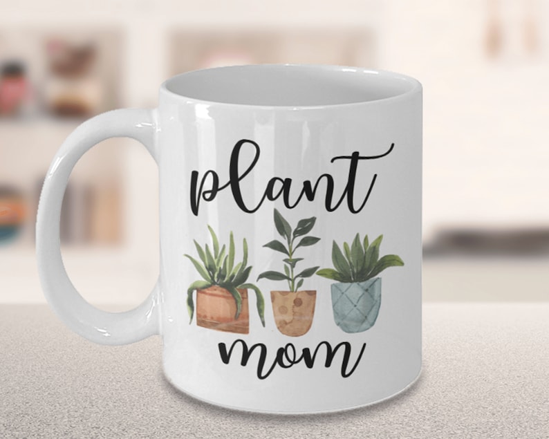 以下が含まれることがあります： 白いセラミック製のマグカップに、黒い筆記体で「plant mom」と書いてあり、緑の葉のついた鉢植えの植物が3つ描かれています。