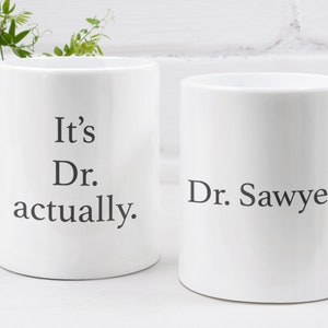 May include: Two white ceramic mugs with black text. The mug on the left says "It's Dr. actually." The mug on the right says "Dr. Sawyer."