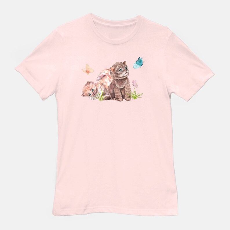 May include: A pink t-shirt with a watercolor illustration of two kittens looking up at a blue butterfly. The kittens are brown and white with pink flowers and green grass around them.