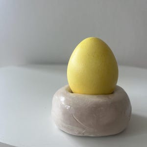 Könnte beinhalten: Ein gelbes, eiförmiges Objekt liegt in einem beigen, abgerundeten Keramikhalter. Das Ei hat eine strukturierte Oberfläche, und der Halter hat eine glatte, glänzende Oberfläche. Die Komposition ist einfach, mit Fokus auf die kontrastierenden Farben und Texturen.