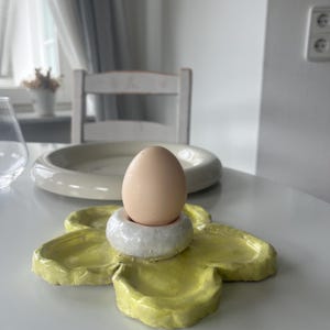 May include: A pale brown egg sits in a white and grey ceramic egg cup, placed on a yellow, flower-shaped ceramic dish. The dish has a glossy finish. A white plate and chair are in the background.
