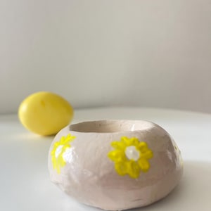 Könnte beinhalten: Eine kleine, runde, beige Keramikschale mit gelben Gänseblümchen-Designs. Im Hintergrund befindet sich ein gelbes, eiförmiges Objekt. Die Schale ist wahrscheinlich handgefertigt und eignet sich für kleine Gegenstände oder als dekoratives Stück.