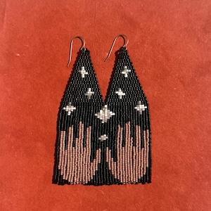 May include: Black beaded earrings with a celestial design. The earrings feature a black background with white cross-shaped stars and pink and white beaded hand-like shapes. The earrings have silver-colored ear wires.