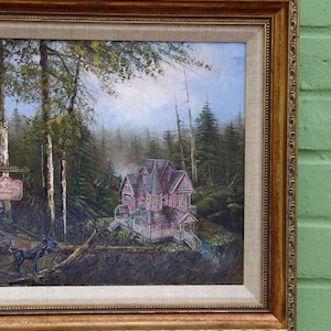 May include: Oil painting in a gold ornate frame depicts a pink Victorian house nestled in a forest. The artwork features a sign, a black creature, and a detailed landscape with trees and a misty background.