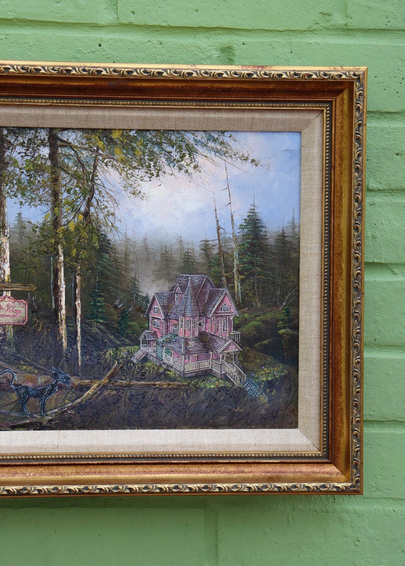 May include: Oil painting in a gold ornate frame depicts a pink Victorian house nestled in a forest. The artwork features a sign, a black creature, and a detailed landscape with trees and a misty background.
