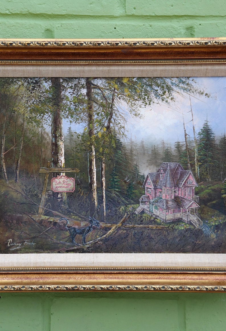 May include: Oil painting of a pink Victorian house nestled in a forest, with a sign that reads "Lake Rose Cottage". The artwork features a detailed landscape with trees, a misty background, and a decorative gold frame.
