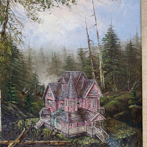 May include: Oil painting of a pink Victorian house nestled in a forest. The house has multiple gables and a wraparound porch. The artwork is framed with a neutral linen-textured border, and the background features tall evergreen trees.