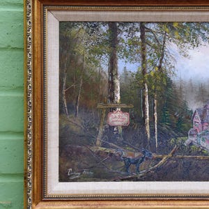 May include: A framed landscape painting with a gold ornate frame. The artwork depicts a forest scene with trees, a sign, and a house. A black sculpture of an animal is in the foreground. The painting is signed by the artist.