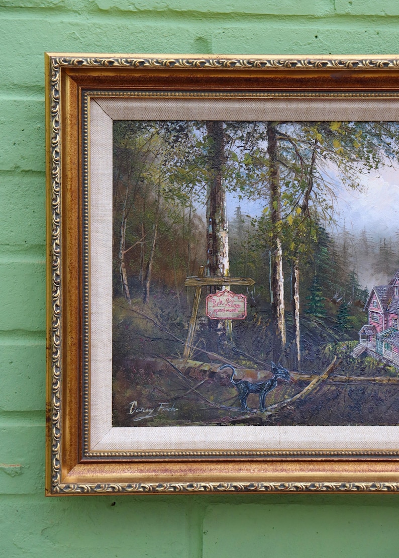 May include: A framed landscape painting with a gold ornate frame. The artwork depicts a forest scene with trees, a sign, and a house. A black sculpture of an animal is in the foreground. The painting is signed by the artist.