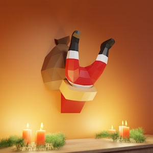 Może przedstawiać: Postać Świętego Mikołaja w kolorze czerwonym i białym jest uwięziona do góry nogami w brązowym worku. Postać jest wykonana z papieru i ma trójwymiarowy design. Postać wisi na ścianie z ciepłym pomarańczowym tłem. Przed postacią stoją trzy zapalone świece.