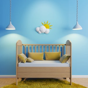 Puede incluir: Una cuna de madera con un móvil de papel en forma de nube y sol blanco y amarillo colgando sobre ella. La cuna tiene un colchón blanco y almohadas amarillas. La cuna está frente a una pared azul claro.