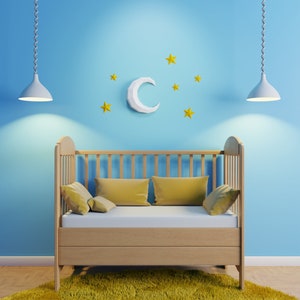 Puede incluir: Una cuna de madera con una luna de papel blanco y estrellas amarillas en una pared azul claro. La cuna tiene un colchón blanco y almohadas amarillas.