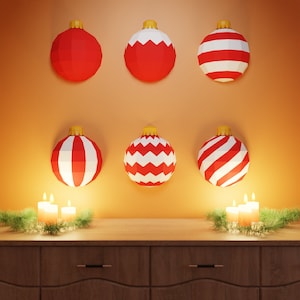 May include: Six red and white Christmas ornaments with a chevron or striped pattern hang on a wall with two lit candles on either side of them.