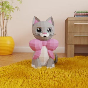 Op de afbeelding: Een grijze en witte origami-kat met een roze strik, zittend op een geel pluche kleed.