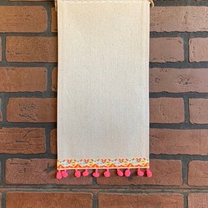 May include: A rectangular wall hanging made of off-white fabric, with a wooden dowel at the top and a decorative trim at the bottom. The trim features a colorful embroidered design with pink pom-poms. A jute rope is used for hanging.
