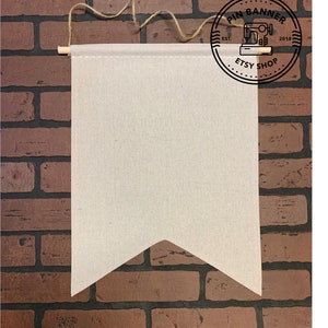 Puede incluir: Una pancarta de lona beige en blanco con una varilla de madera en la parte superior y una cuerda marrón que cuelga de ella. La pancarta tiene forma de banderín con la parte inferior puntiaguda. La pancarta está colgada contra una pared de ladrillo. Un logotipo circular es visible en la esquina superior derecha de la imagen. El logotipo dice "Pin Banner Etsy Shop Est. 2019" con un icono de máquina de coser en el centro.