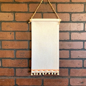 May include: A rectangular, off-white fabric wall hanging with a decorative trim featuring colorful geometric patterns and small, cream-colored pom-poms. The hanging is suspended by a natural rope and wooden dowel.