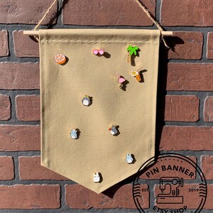Puede incluir: Un banner de lona beige con una varilla de madera en la parte superior, colgado de una pared de ladrillo. El banner tiene una variedad de pines de esmalte unidos a él, incluyendo una palmera, un flamenco, una bebida con una rodaja de naranja, un corazón, una rodaja de naranja y varios personajes de dibujos animados. El banner tiene un círculo blanco y negro en la parte inferior con el texto "PIN BANNER EST 2019 ETSY SHOP".