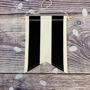 Puede incluir: Un banderín de tela a rayas blancas y negras con la parte inferior puntiaguda. El banderín está colgado de una cuerda atada a dos listones de madera.