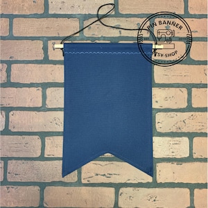 May include: A dark blue fabric banner with a scalloped edge and a pointed bottom. The banner is hanging from a wooden dowel with a string. The banner is blank and ready to be customized.
