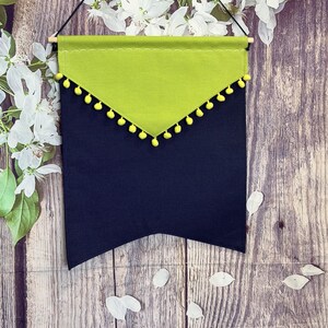 Puede incluir: Un banderín decorativo para colgar en la pared con una base azul marino y una sección superior verde lima. La sección verde está adornada con pompones amarillos. El banderín está suspendido de una varilla de madera con una cuerda negra.