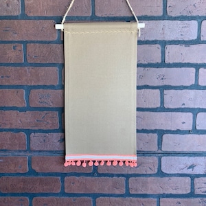 May include: A beige fabric banner with a white and pink trim and a row of pink pom-poms along the bottom edge. The banner is hanging from a brick wall.